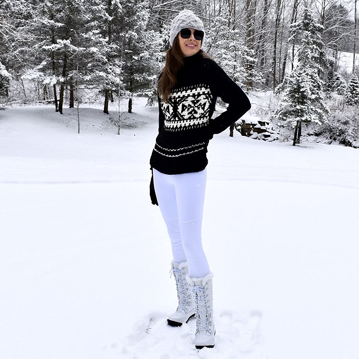 ski winter outfit with white leggings, a black Christmas sweater, a beanie, aviator glasses, and white boots